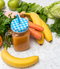 Freshly prepared smoothie in a jar with a straw. There are carrots, bananas, oranges, and greens nearby. Selective focus.
