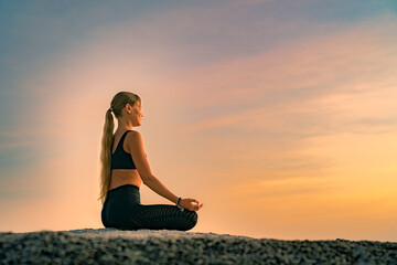 Young caucasian woman with long hair meditating in a black clothes for yoga on a rocks on the top. On the background  colorful sunrise sky.