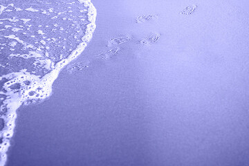 Toned with Very Peri color Kid footprints on sand at the Beach and cooming foamy wave. Smooth costline. Horizontal with copy space. Travelling and holiday concept