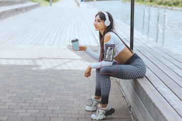 A woman in sportswear listens to music on headphones after a workout and drinks coffee on the river...