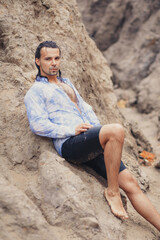 A man with dark long hair sits on a sandbank in a wet shirt and wet hair