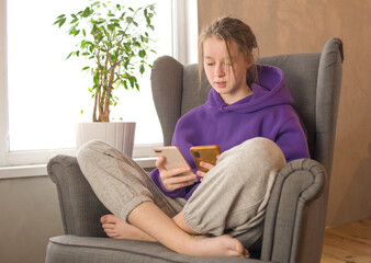 Girl in a lilac sweater sits with a phone in a chair near the window. Girl relaxing in chair using two smartphones.Communication, study at home, mobile apps, lockdown, technology, lifestyle, online