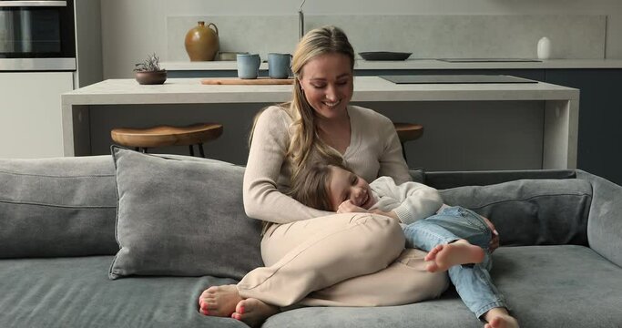 Beautiful young loving mother tickles her little daughter. Preschool 5s girl enjoy playtime with mom, play on sofa at modern home. Family leisure, active games, parent and kid time together concept
