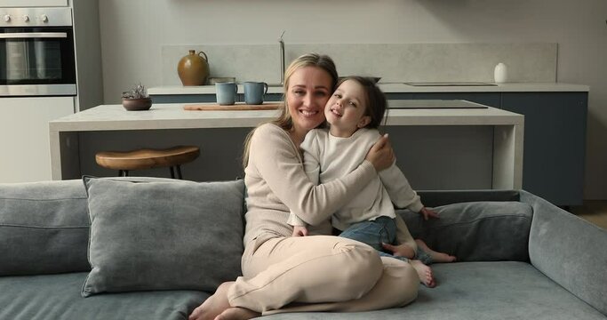 Happy motherhood, wellbeing family portrait concept. Blond woman her little 4s preschool daughter hugging sit on sofa smile look at camera, mother feeling unconditional love, kid enjoy tender moments