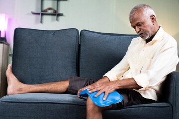 senior man using Hot water massage bag for knee joint pain relief while sitting on sofa at home - concept of natural hot water treatment, muscle relaxtion and osteoarthritis. - obrazy, fototapety, plakaty