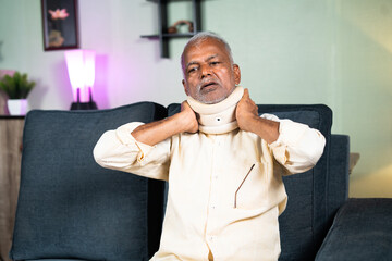 Indian senior old man using neck brace or carvical collars due to neck pain at home - concept of...