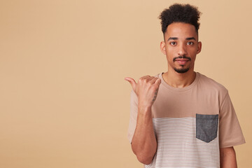 Studio portrait of young african american male points aside at copy space with excited, shocked facial expression. posing over beige background
