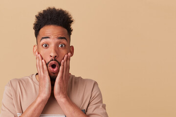 Studio portrait of young african american male looking directly into camera with. shocked, surprised facial expression, touching his cheek. posing over beige background