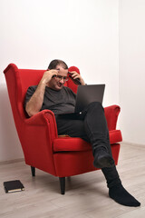 Middle-aged man in glasses with a laptop on his lap and telephones sitting in a red armchair. mockup