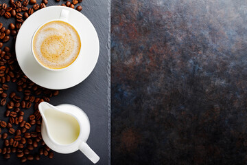 Coffee and roasted coffee beans on a slate board. Cup of coffee on a white saucer and a milk jug on a dark background. Copy space. Top view