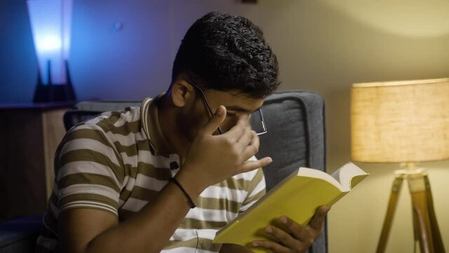 Young student with spectacles rubbing eyes due to eyes strain while reading book during late night - concept of feeling tired, early morning study and education.