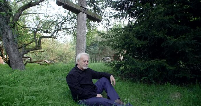 Sad old gray haired man sits on the grave with big old cross at the cemetery, sorrow and sadness about lost of close people, 4k 60p Prores