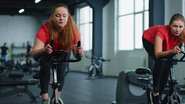 Group of smiling friends at gym exercising on stationary bike. Happy cheerful athletes training on exercise bike. Young women working out at class in gym. Weight loss, fat burning. Modern active sport