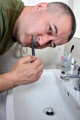 A white man is brushing his teeth in the bathtub above the sink. He looks into the camera. The tap is open and water is flowing.