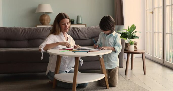 Happy young single mom little son friends sit on heated floor on carpet by small table draw funny pencil sketches together. Affectionate foster mum watch adopted kid boy preschooler creating picture