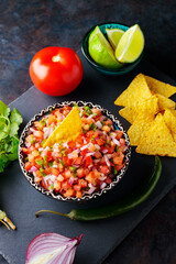 Bowl of tasty Pico de Gallo salsa on dark background. Pico de Gallo salsa and ingredients. Mexican food concept. Top view
