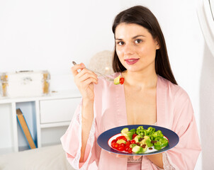 Obraz na płótnie Canvas Positive pretty woman in bathrobe holding fork and eating vegetable salad in bed at home