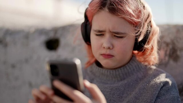 cute teen sad sitting outside on a sunny day and listening to music in wireless headphones via the Internet on a mobile phone