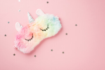 Top view photo of funny multicolored fluffy unicorn sleeping mask and star shaped confetti on isolated pastel pink background with copyspace