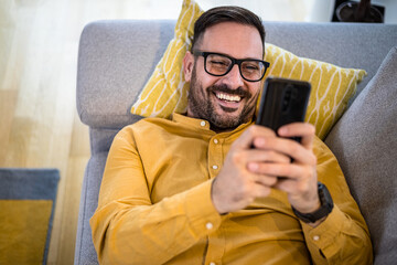 Cheerful young adult mature man happy smiling using smart phone resting lying down on sofa couch at...
