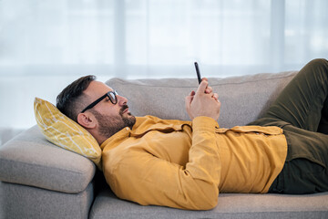 Young adult mature man using smart phone resting lying down on sofa couch at home messaging on mobile phone Guy with eyeglasses relaxing at home surfing social media buying purchasing paying ordering