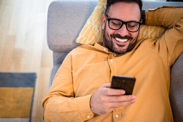 Cheerful young adult mature man happy smiling using smart phone resting lying down on sofa couch at...
