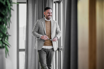 Obraz na płótnie Canvas Portrait of young adult smiling cheerful businessman entrepreneur manager in casuals holding glasses standing next to the window in modern office looking away satisfied and successful.