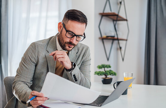 Serious Pensive Thoughtful Focused Young Adult Casual Businessman In Office Working With Laptop Computer Papers And Documents Looking At Paper Report Graphs Chart Diagram Spreadsheet