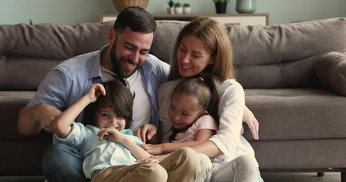 Bonding affectionate family couple custodians with little adopted kids tickling on warm floor playing with sincere laughter having fun. Adorable playful children boy girl messing kidding with mom dad