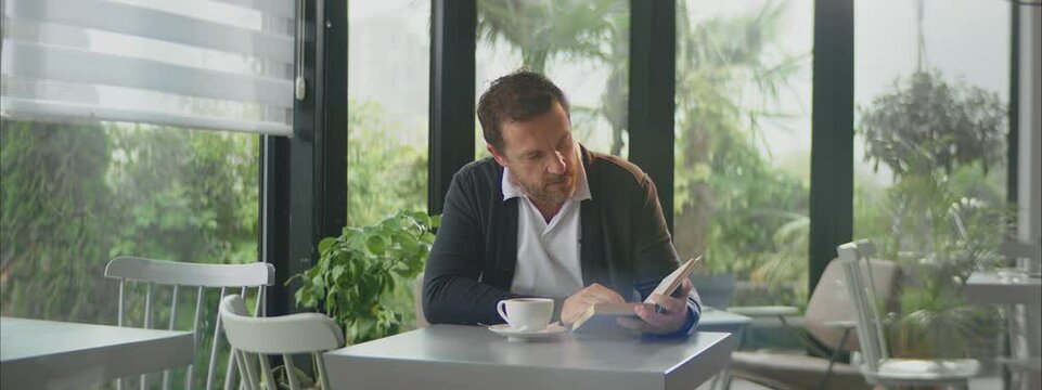 Mid adult Caucasian man reading book while sitting at small table in cafe. Waiting for client. Entrepreneur working outside office.