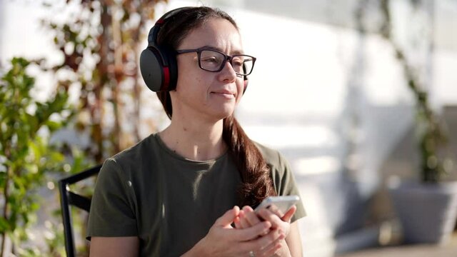An attractive adult woman in stylish glasses and wireless headphones listens to a radio program in a cafe
