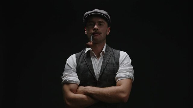 Portrait of young handsome confident rude mustachioed man in vintage suit, cap looking at camera, kneads his hands and standing arms crossed on black background. Mens power promotion theme template