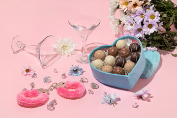 Valentines day creative layout with heart shaped chocolate box, flowers bouquet, martini glasses and furry handcuffs on pastel pink background. 80s or 90s retro fashion aesthetic love concept. 
