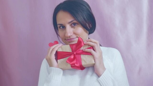 Smiling funny young brunette in a white sweater with a gift and a red heart is dancing on a pink background. Valentine's Day