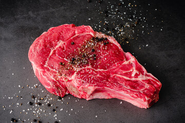 Raw Bone-In Ribeye Steak Seasoned with Kosher Salt and Black Pepper: An uncooked bone-in beef steak sprinkled with salt and pepper on a dark background