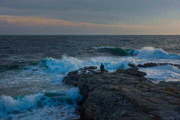 Man sea storm beach. The element large waves break on the stone shore. Seascape with an impending storm. A man poses against the background of the raging surf. Resort concept, climate weather changes