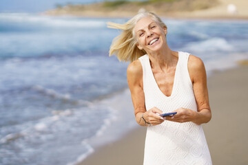 Smiling mature woman walking on the beach using a smartphone.