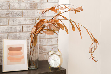 Vase with autumn branches, photo frame and alarm clock on table near light brick wall