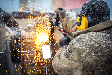 Worker in dirty clothes saws metal in winter in snowfall