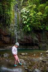 Traveler girl wearing sport jacket walking on shores of waterfall. Travel lifestyle. View from back.