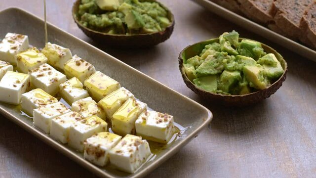 Pouring olive oil on spiced feta cheese cubes snack. Served with chunky guacamole and bread.