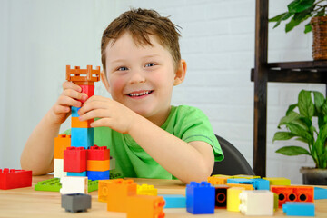 The preschooler sits at the table and plays with cubes, constructor. He looks into the frame, smiling. Horizontal photo. Multi-colored cubes on the table.