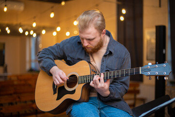 Obraz premium A young guy with a beard plays an acoustic guitar in a room with warm lighting