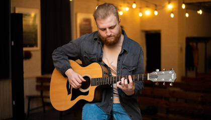 Obraz premium A young guy with a beard plays an acoustic guitar in a room with warm lighting