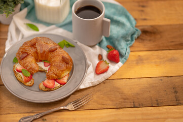 On a white earthenware plate croissant with fresh strawberries, almonds and mint leaves. Cup of coffee and milk in a jug. There is a fork nearby. Wooden background. Culinary blog, menu design.