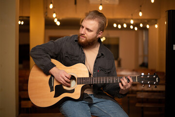 A young guy with a beard plays an acoustic guitar in a room with warm lighting