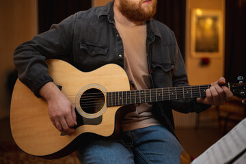 Obraz premium A young guy with a beard plays an acoustic guitar in a room with warm lighting