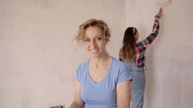 Woman middle aged looking at the camera. Woman friends making repair together, painting white wall in empty apartment with rollers, slow motion
