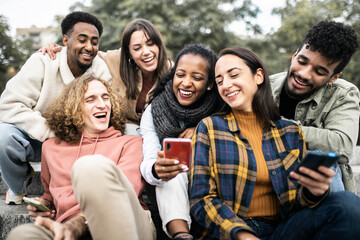 Happy group of young people having fun sharing video content on social media platform - Millennial multiracial friends using smartphone while sitting outdoors - Youth and technology concept