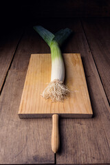 A leek resting on a wooden chopping board on a rustic wooden table in a dark environment.